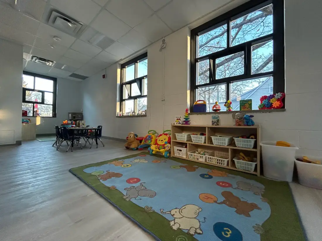 Bright classroom with large windows, colorful rug, and organized toy shelving